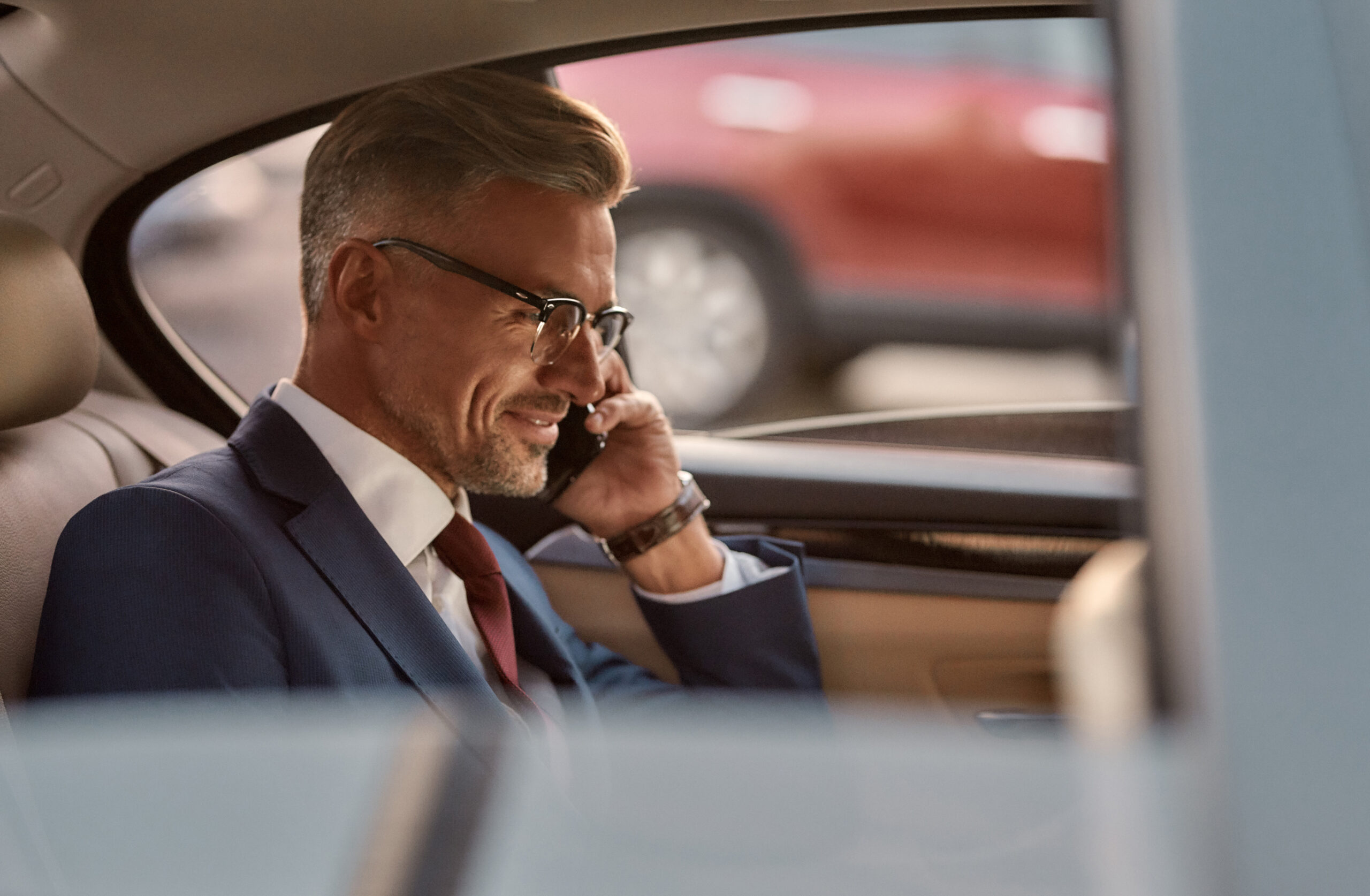 I am always available. Side view of cheerful mature man in classic wear sitting in the car and talking by the phone with client